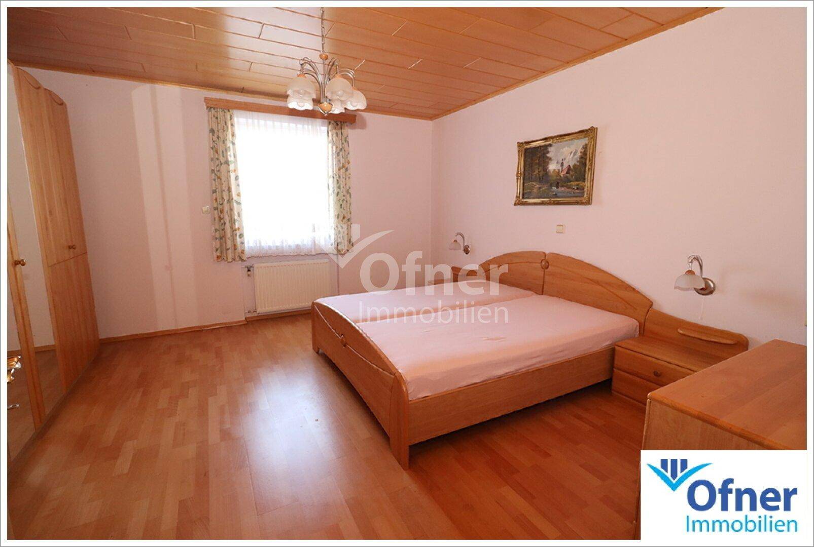 Schlafzimmer mit Holzboden, Doppelbett und Holzmöbeln, hell durch großes Fenster.