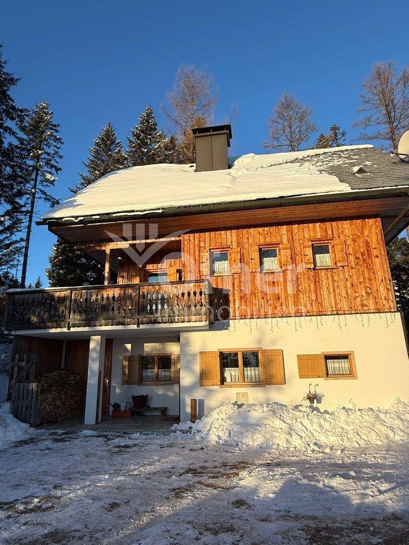 Traditionelles Holzhaus mit verschneitem Dach und Balkon in winterlicher Landschaft.