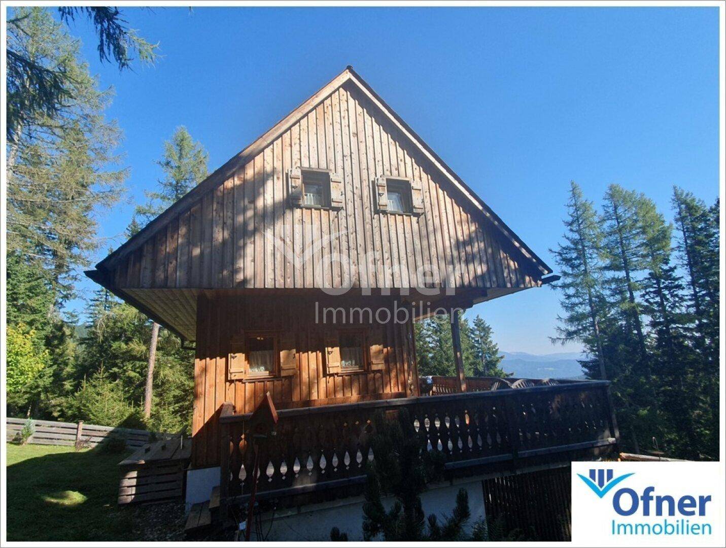 Rustikales Holzhaus mit Satteldach und Balkon, umgeben von grünen Bäumen unter blauem Himmel.