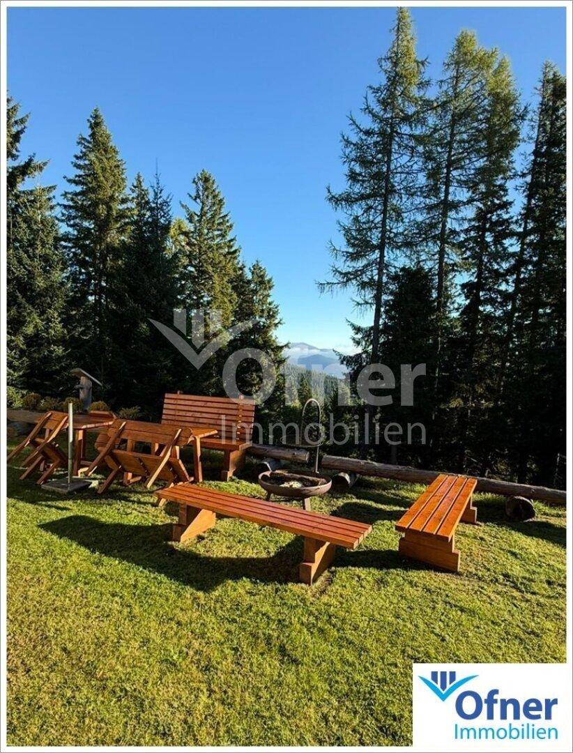 Großer Garten mit Holzbänken, Tischen und Feuerstelle, umgeben von hohen Nadelbäumen.