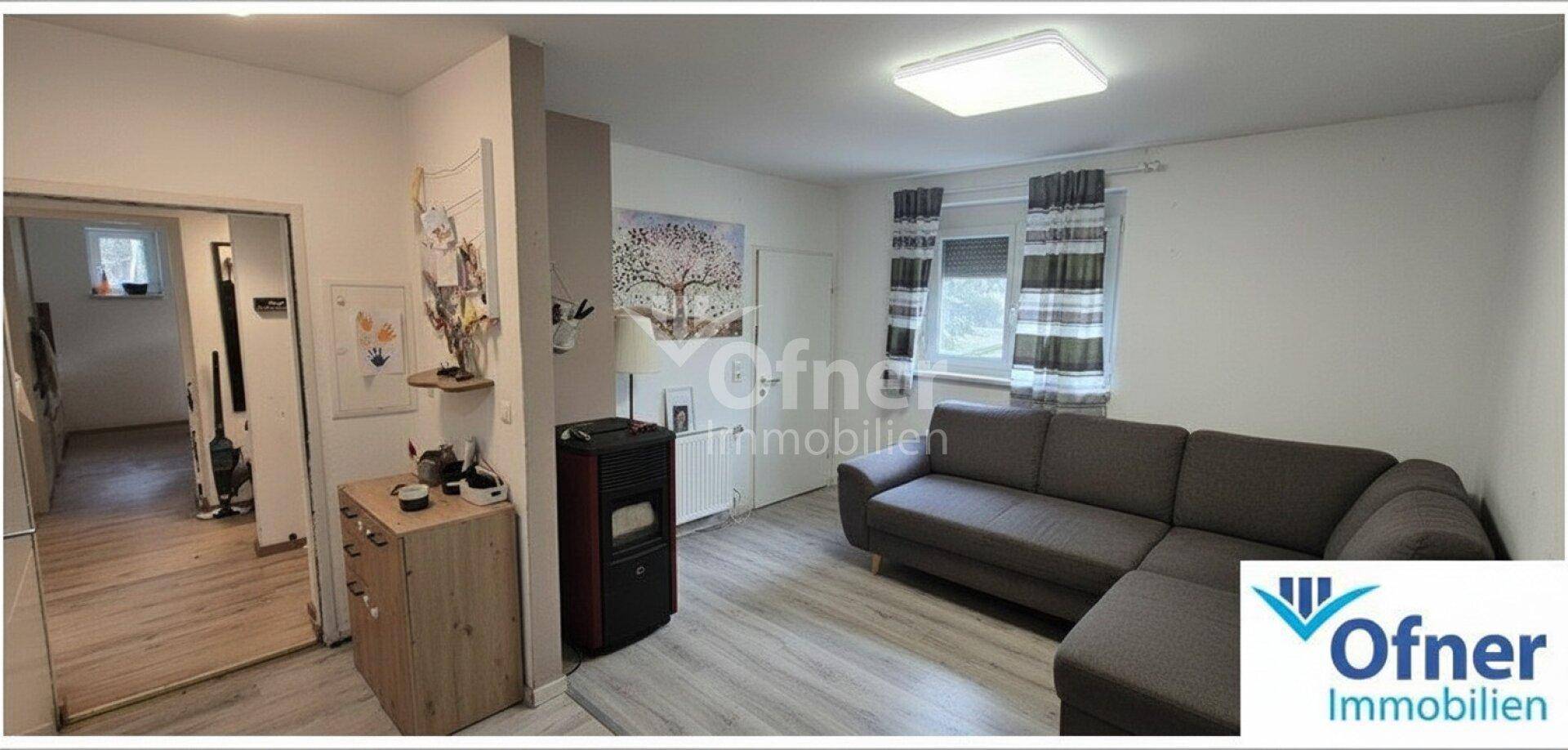 Gemütliches Wohnzimmer mit Sofa, Kaminofen und Laminatboden, hell durch ein Fenster beleuchtet.