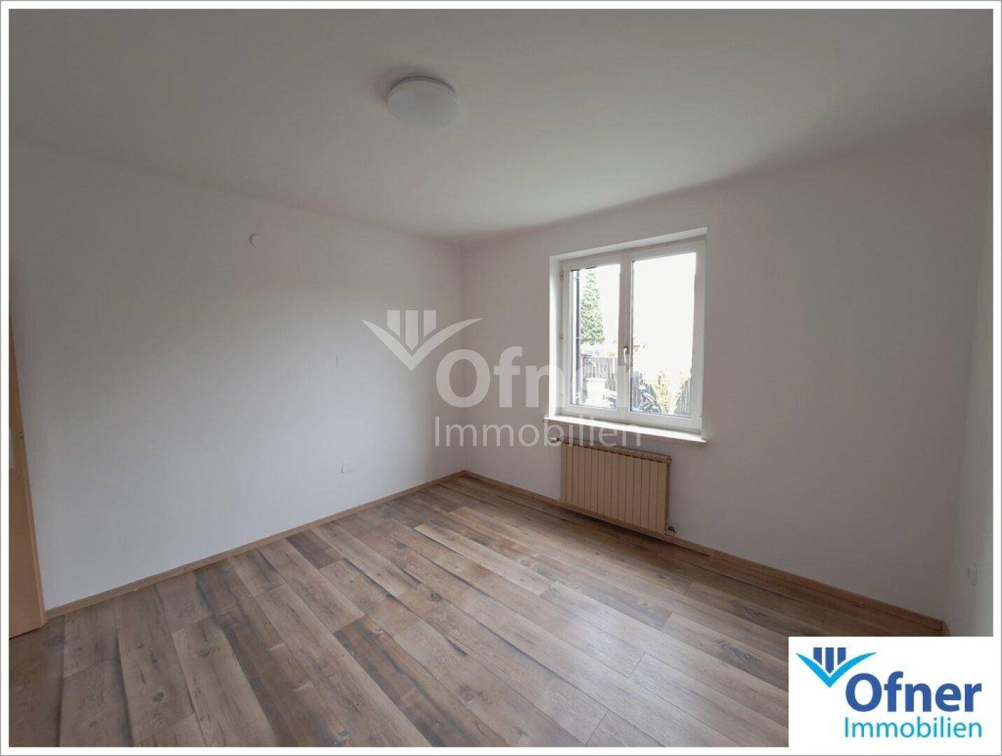 Leeres Zimmer mit Holzboden und einem Fenster, das viel Tageslicht hereinlässt.