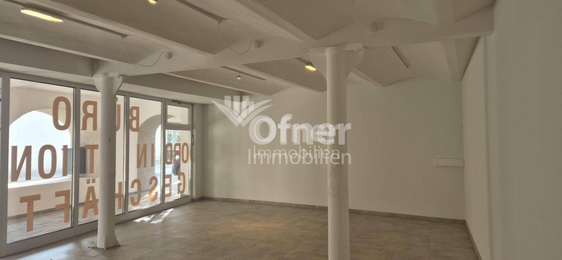 Großer Geschäftsraum mit Glasfront, Fliesenboden und hohen Decken mit sichtbaren Balken.
