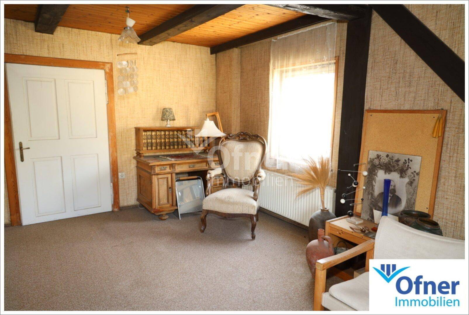 Zimmer mit Teppichboden, Holzbalken an der Decke und einem Schreibtisch am Fenster.
