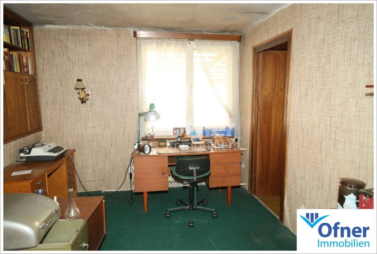 Arbeitszimmer mit Schreibtisch, Bücherregal und Teppichboden, beleuchtet durch ein Fenster.