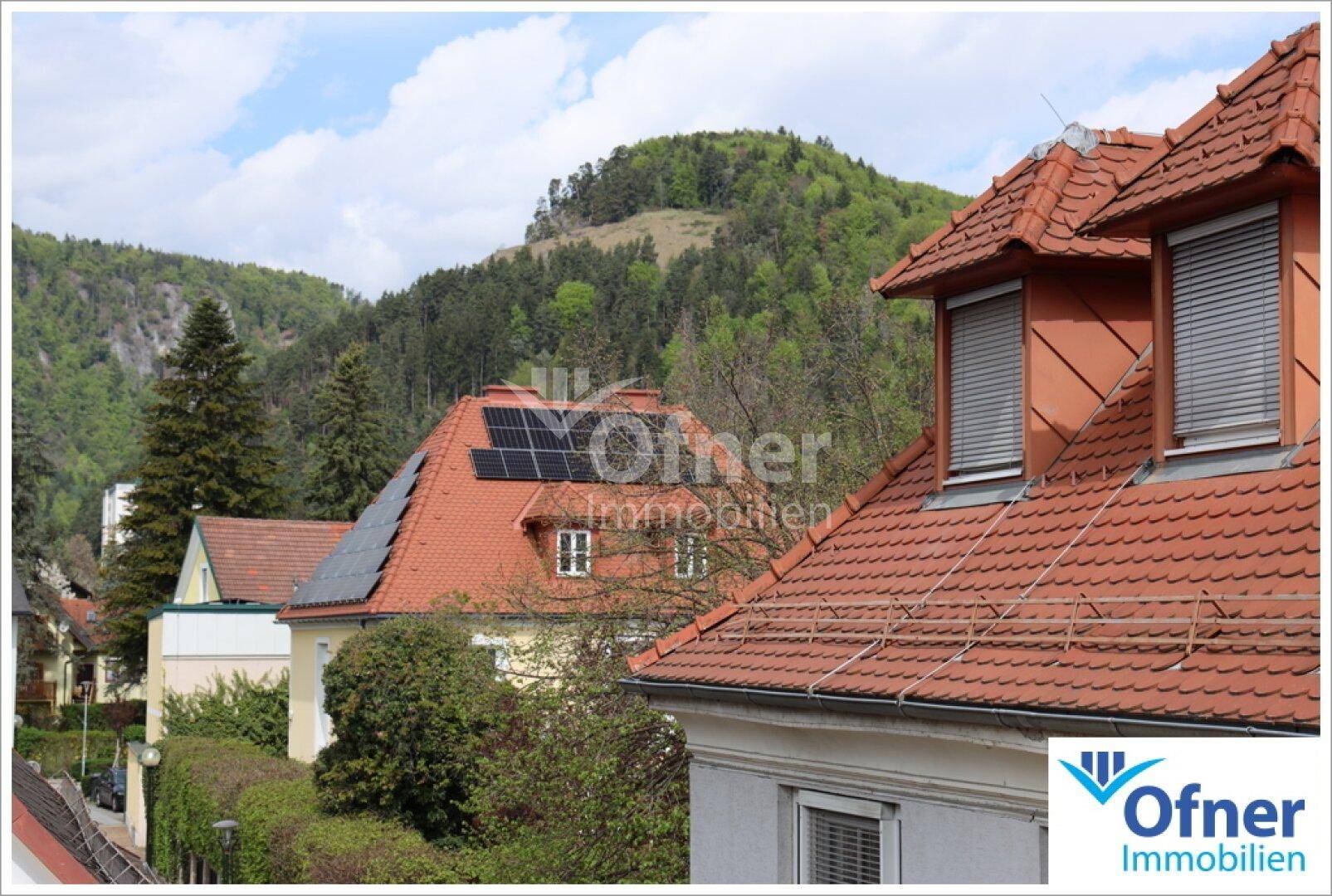 Dächer von Wohnhäusern mit Ziegeln und Photovoltaikanlagen, umgeben von grünen Hügeln.