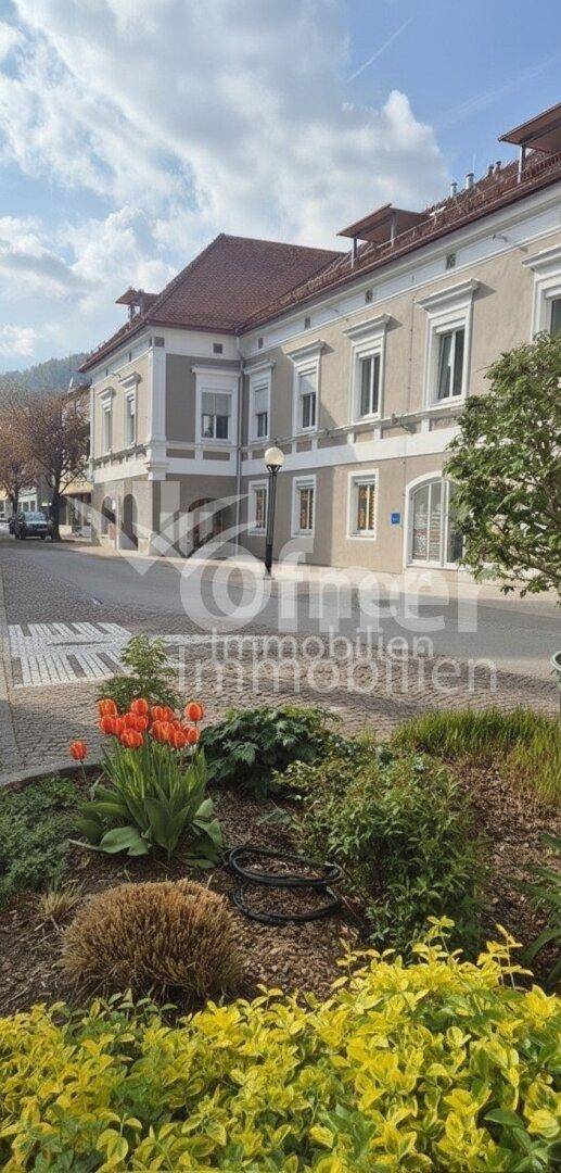 Ansicht des Gebäudes von der Straße mit blühenden Pflanzen und Bäumen im Vordergrund.