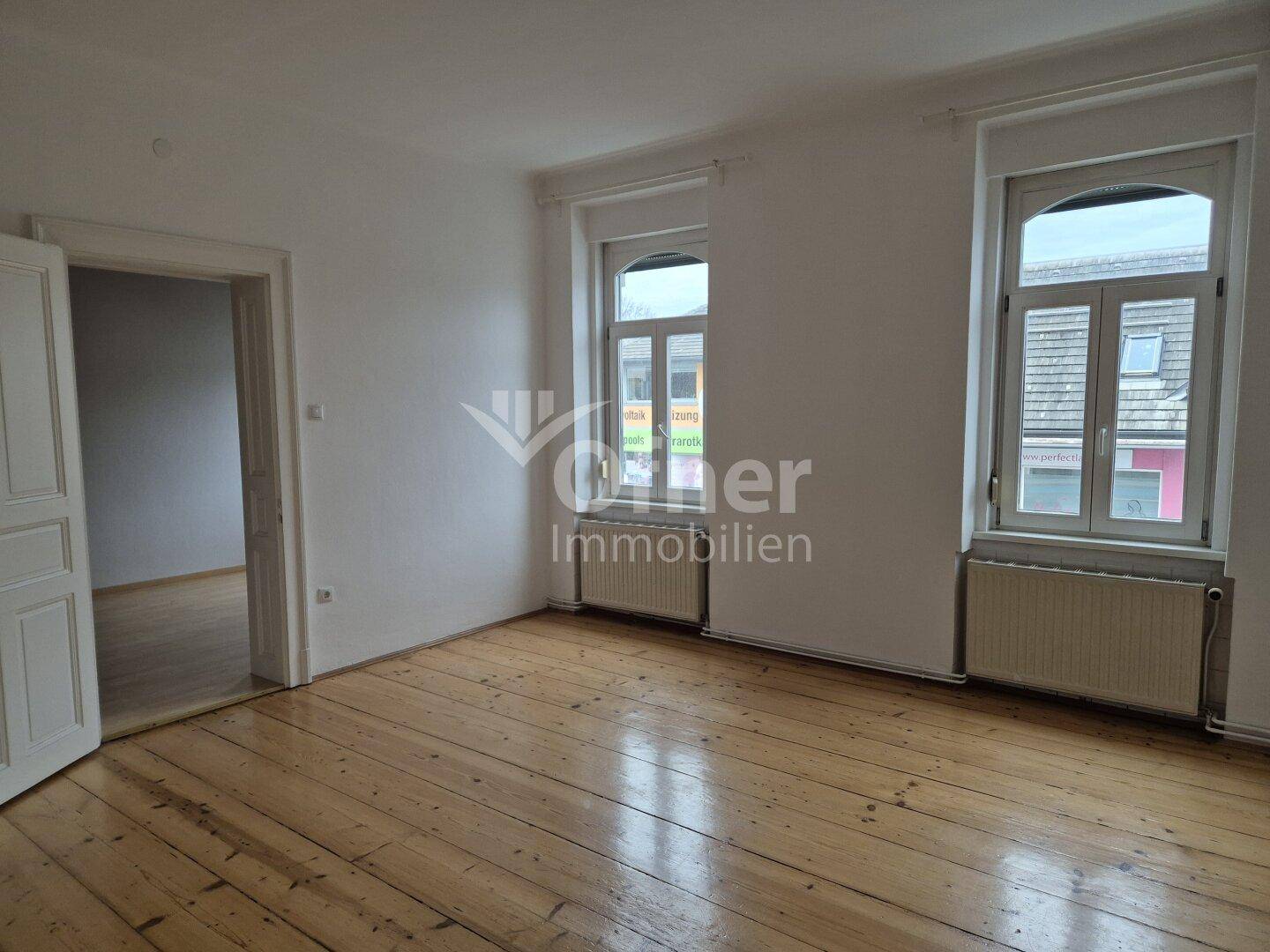 Helles Zimmer mit zwei Fenstern, Holzplankenboden und weiß gestrichenen Wänden.