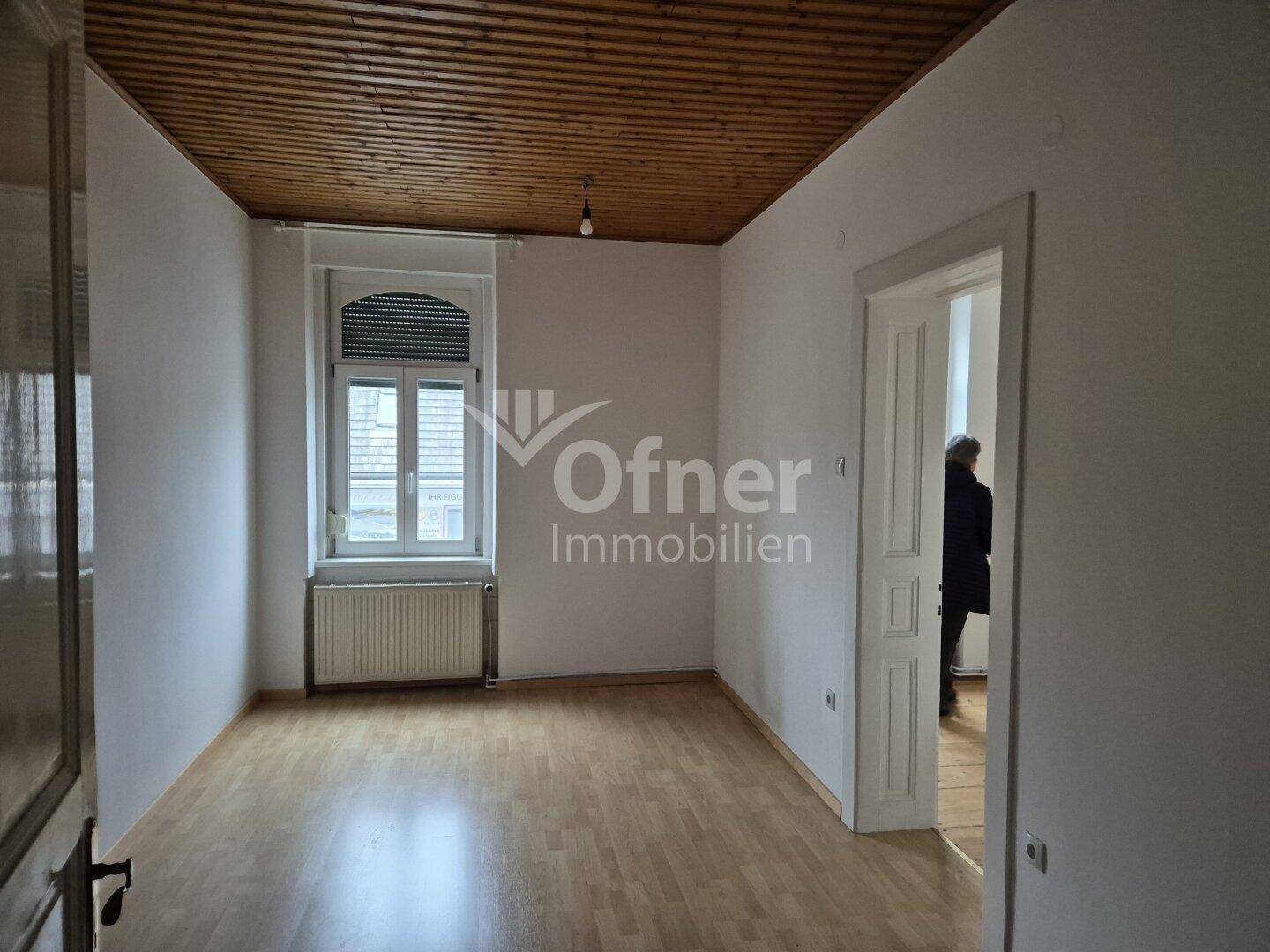 Leeres Zimmer mit hellem Holzparkett, Holzdecke und Fenster mit Heizkörper.