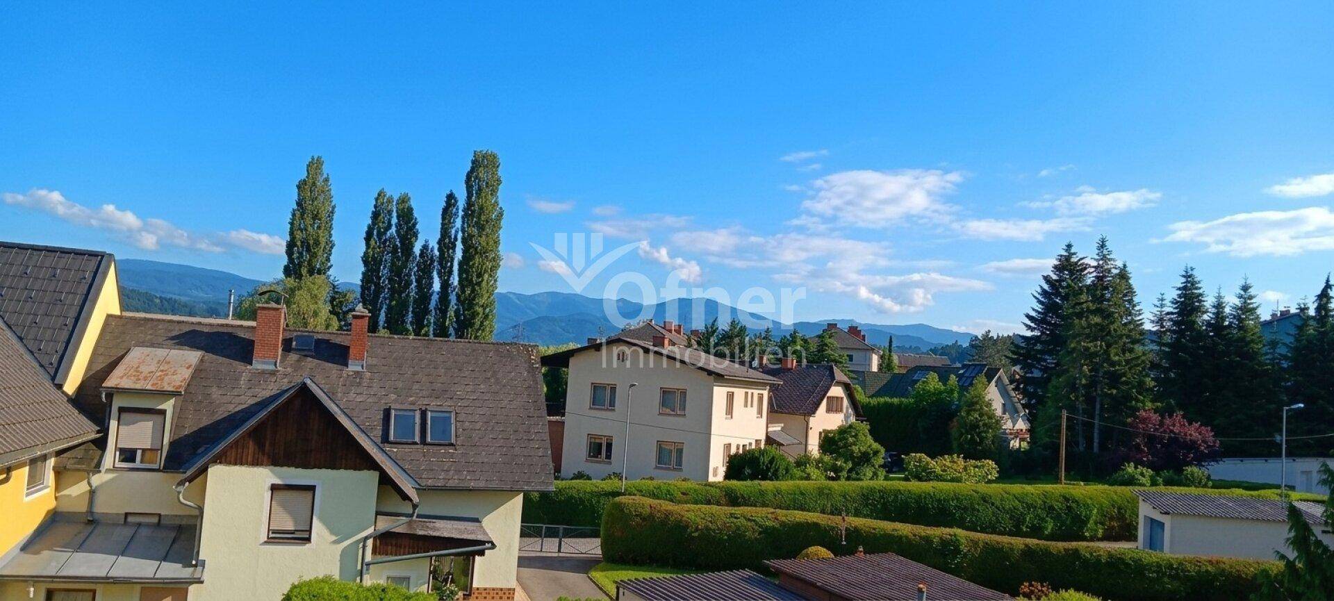 Panoramablick über eine Wohnsiedlung mit Häusern, viel Grün und einer Bergkulisse im Hintergrund.