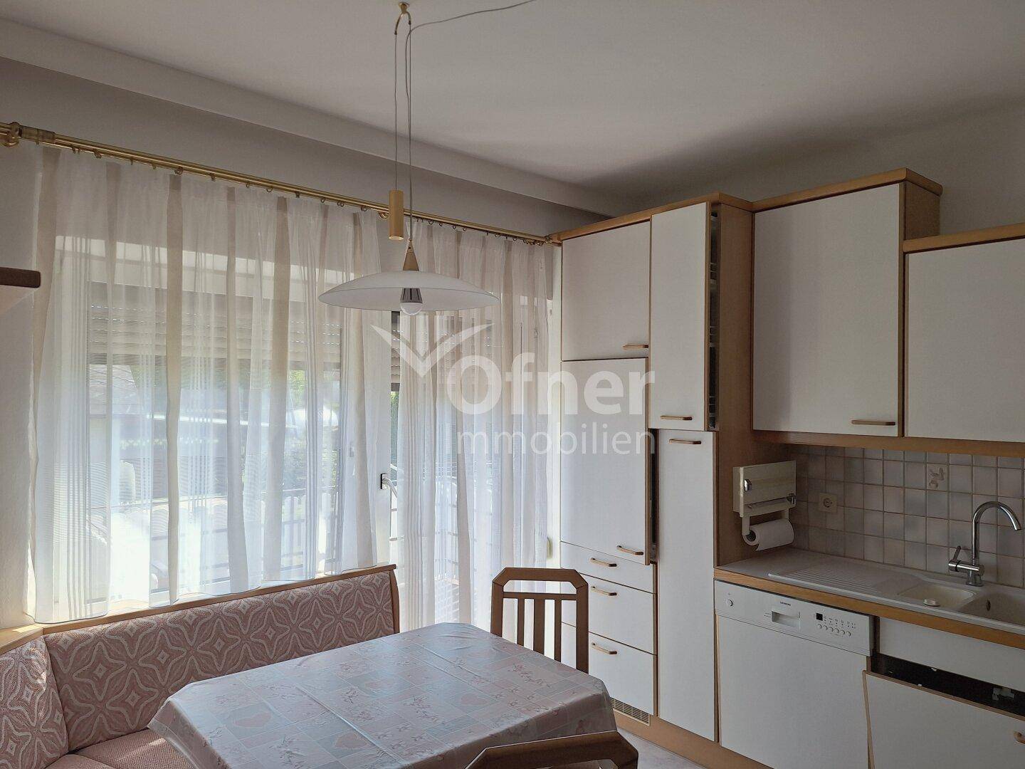 Helle Küche mit weißen Schränken, einer gemütlichen Essecke und großem Fenster mit Vorhängen.