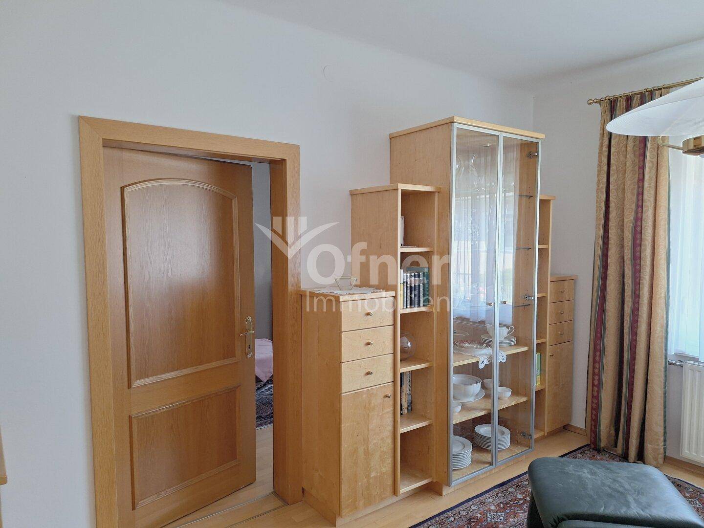 Wohnbereich mit hellen Holzschränken, einer Vitrine und einem Durchgang zu einem weiteren Raum.