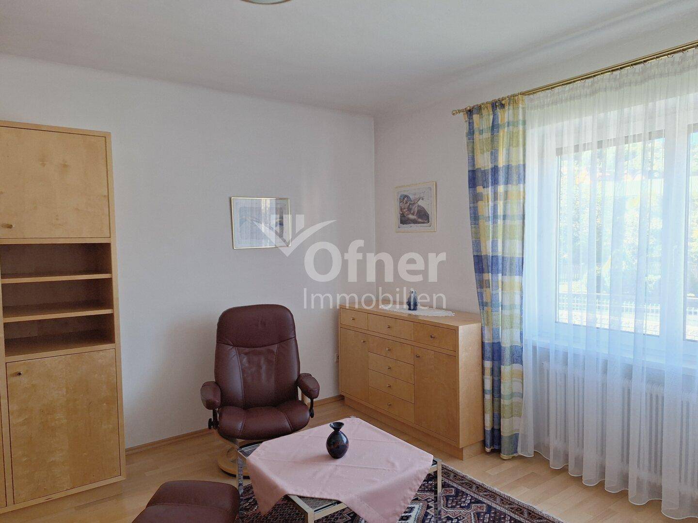 Gemütliches Wohnzimmer mit Holzmöbeln, einem bequemen Sessel und einem großen Fenster für viel Licht.
