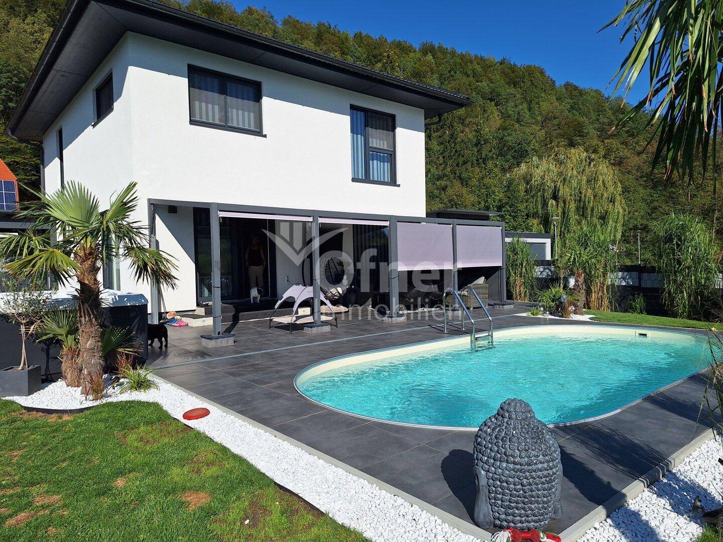 Ansicht des modernen Hauses mit großem Poolbereich, Terrasse und üppiger Vegetation im Hintergrund.