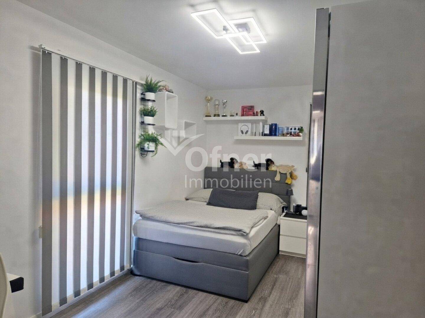 Helles Schlafzimmer mit Einzelbett, modernem Bodenbelag und Fenster mit vertikalen Jalousien.