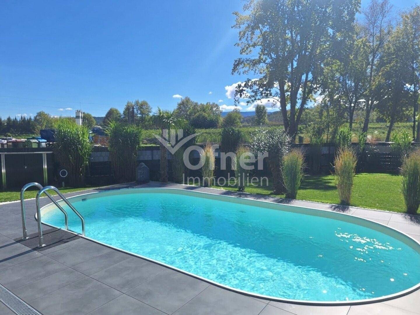 Ovaler Swimmingpool im Garten mit dunklen Fliesen und umgebender Rasenfläche unter blauem Himmel.