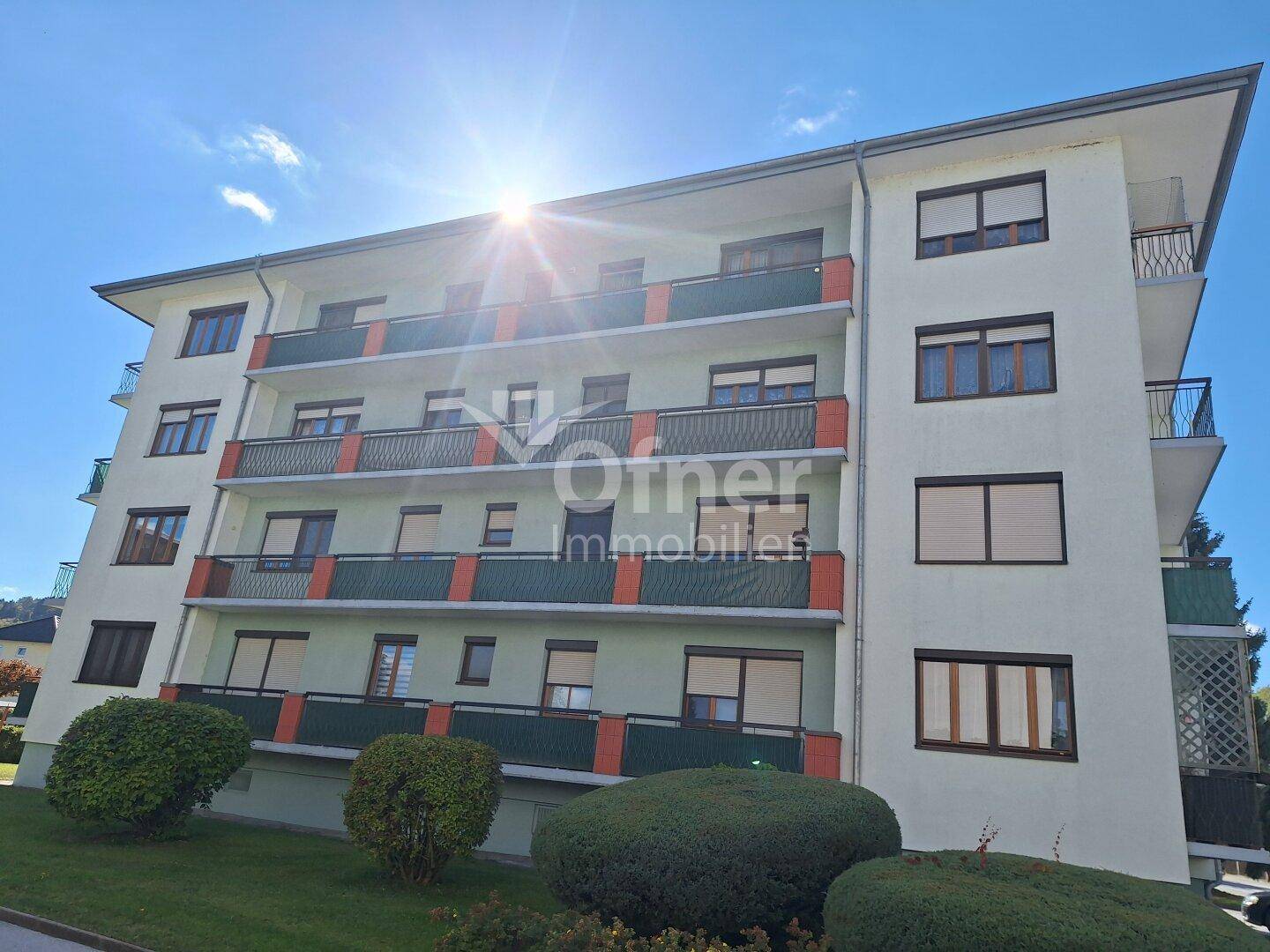 Mehrstöckiges Apartmentgebäude mit Balkonen und grünen Akzenten unter strahlendem Sonnenschein.