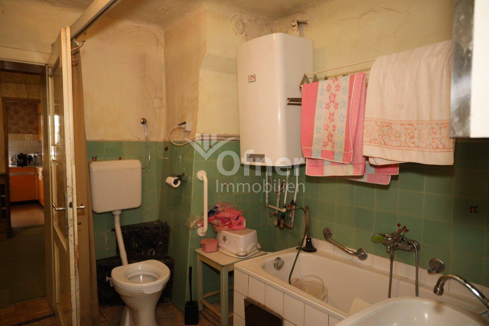 Älteres Badezimmer mit grünen Fliesen, Badewanne, Toilette und Warmwasserboiler.