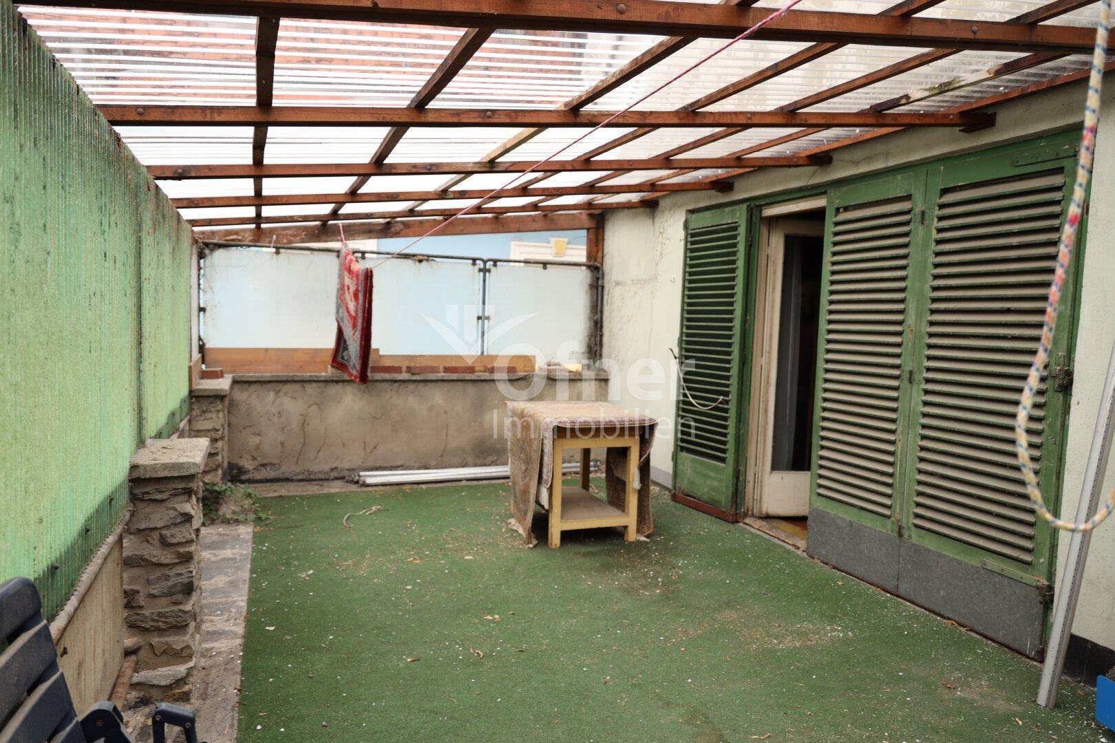 Überdachte Terrasse mit Kunstrasen und grünen Fensterläden.