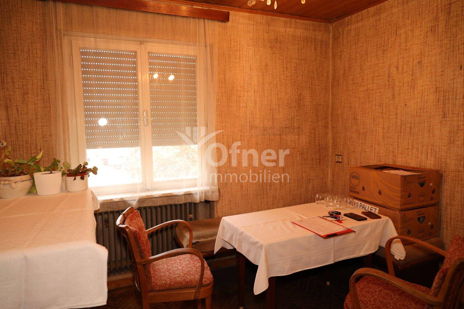 Kleiner Wohnbereich mit Fenster, Rollos und einfacher Möblierung.