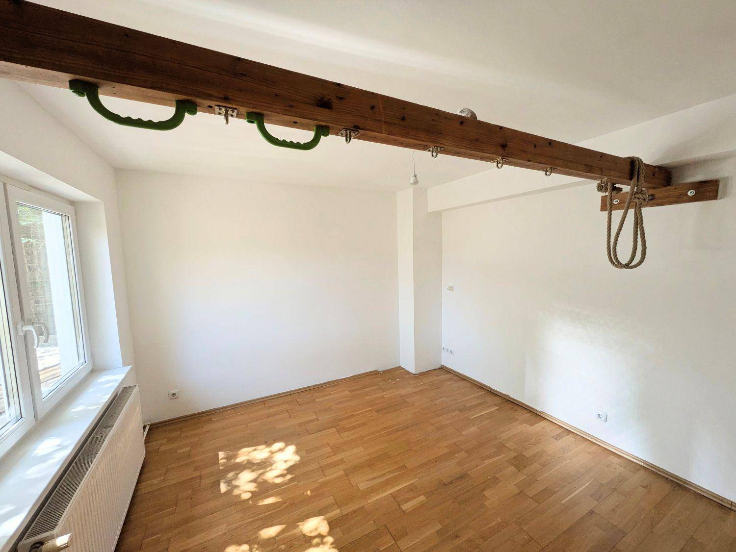 Zimmer mit Parkettboden, Fenster und einem sichtbaren Holzbalken mit Klettergriffen.