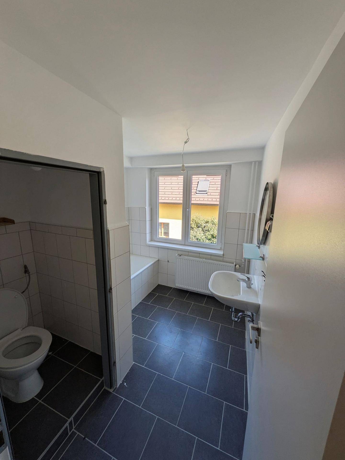 Badezimmer mit dunklen Bodenfliesen, weißen Wandfliesen, Badewanne und Fenster.