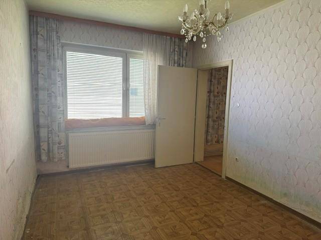 Helles Zimmer mit Parkettboden, gemusterter Tapete und Zugang zu einem weiteren Raum.