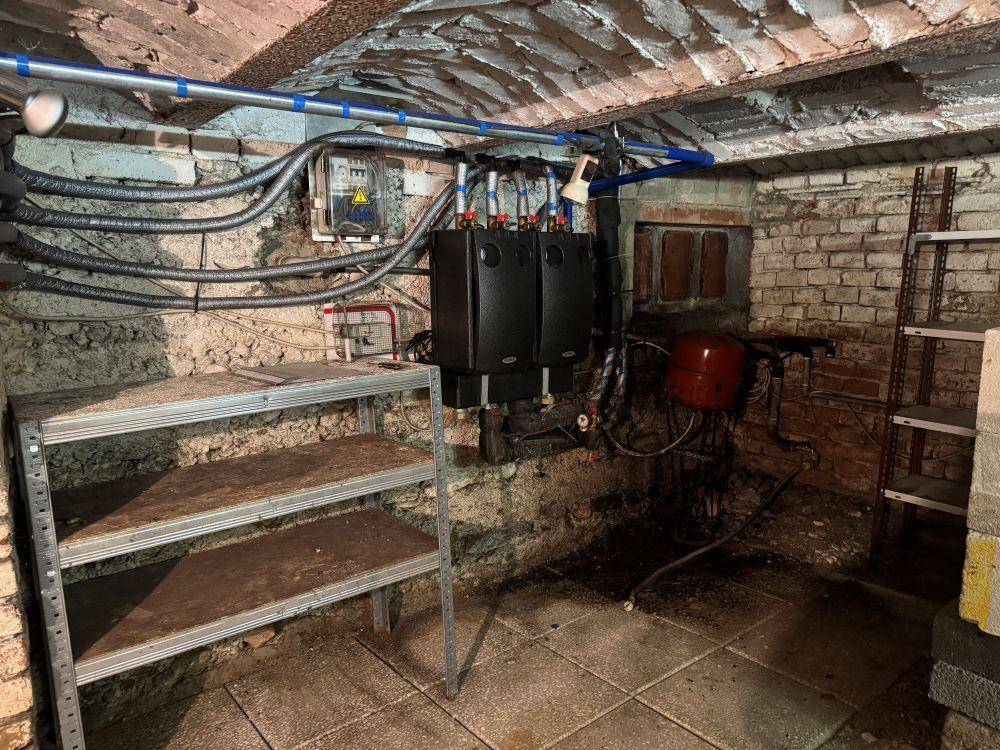 Kellerraum mit Heizungsanlage, Rohren und Regalen, der als Technik- oder Abstellraum dient.
