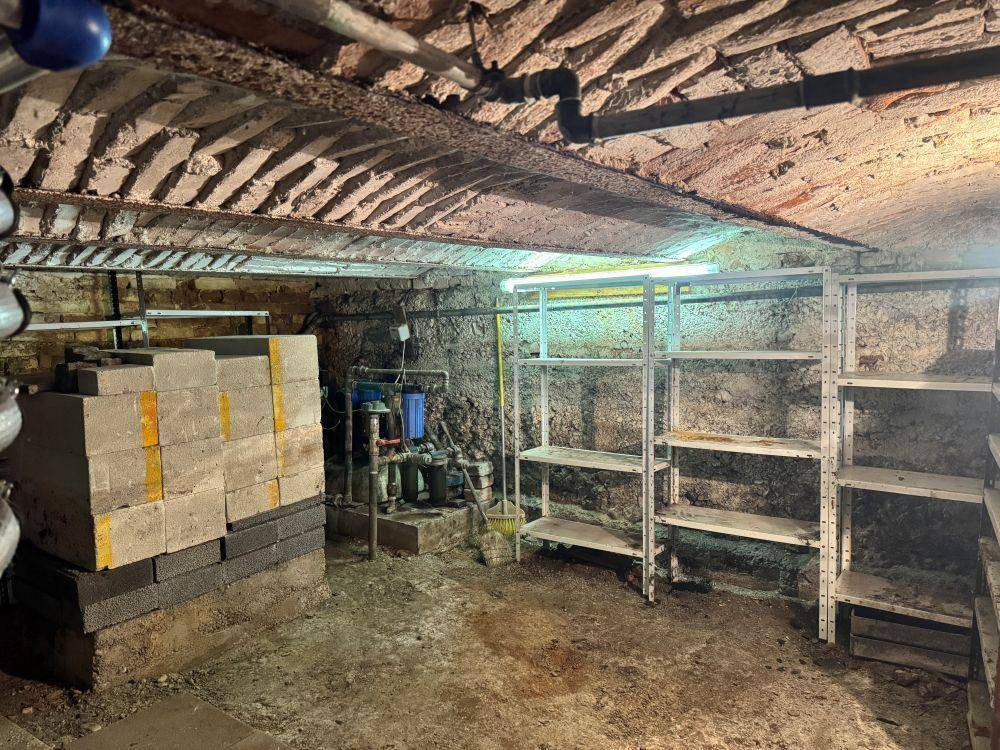 Kellerraum mit sichtbaren Rohren, Steinwänden und Regalen, der als Technik- oder Abstellraum dient.