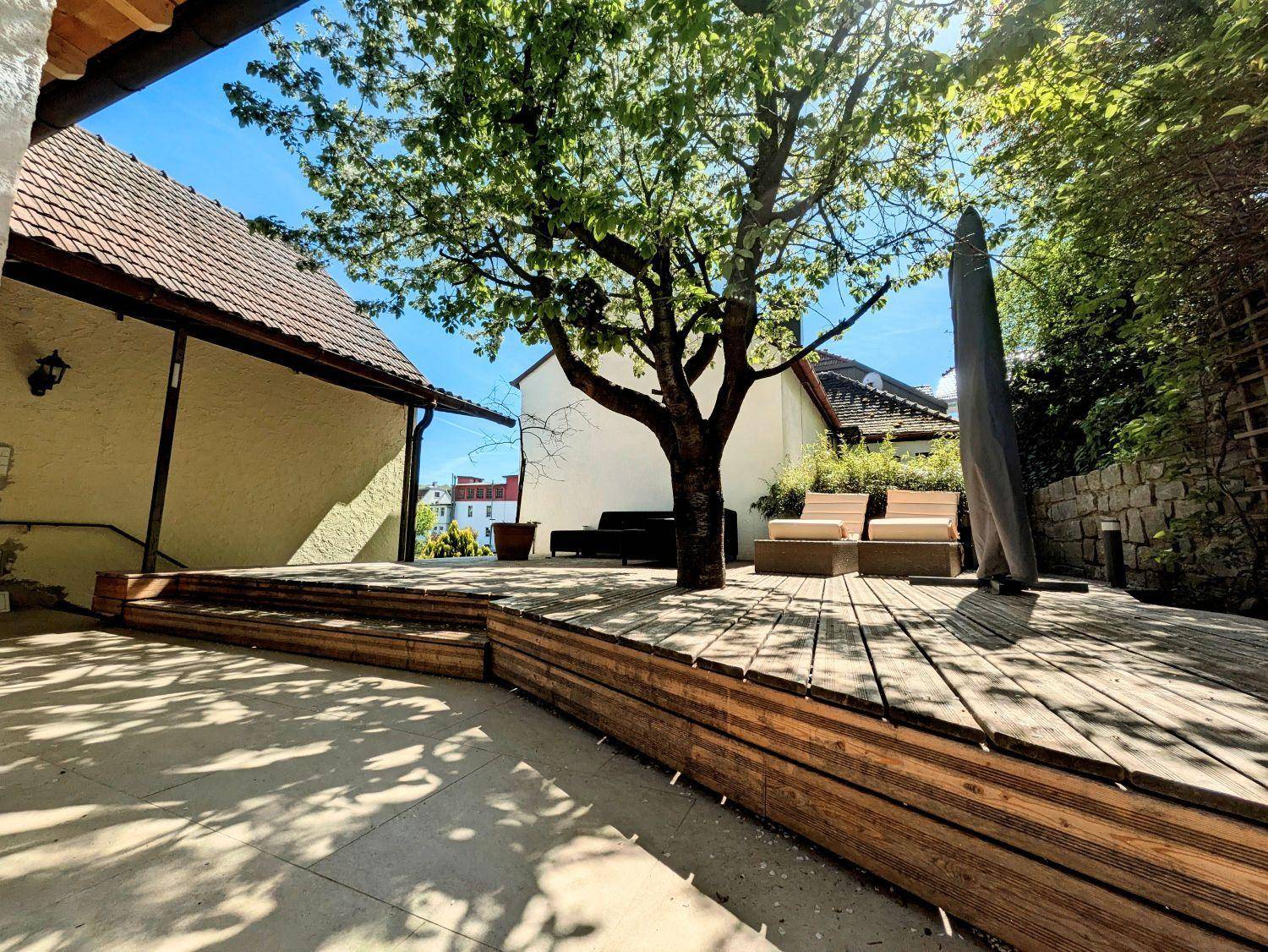 Geräumige Holzterrasse mit Lounge-Möbeln und einem großen Baum als Schattenspender.