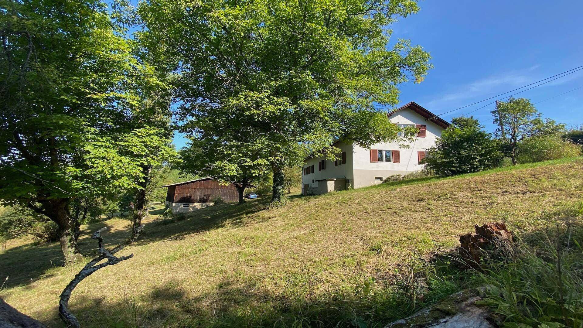 Ländliches Haus auf einem Hügel mit viel Grünfläche und Bäumen unter blauem Himmel.