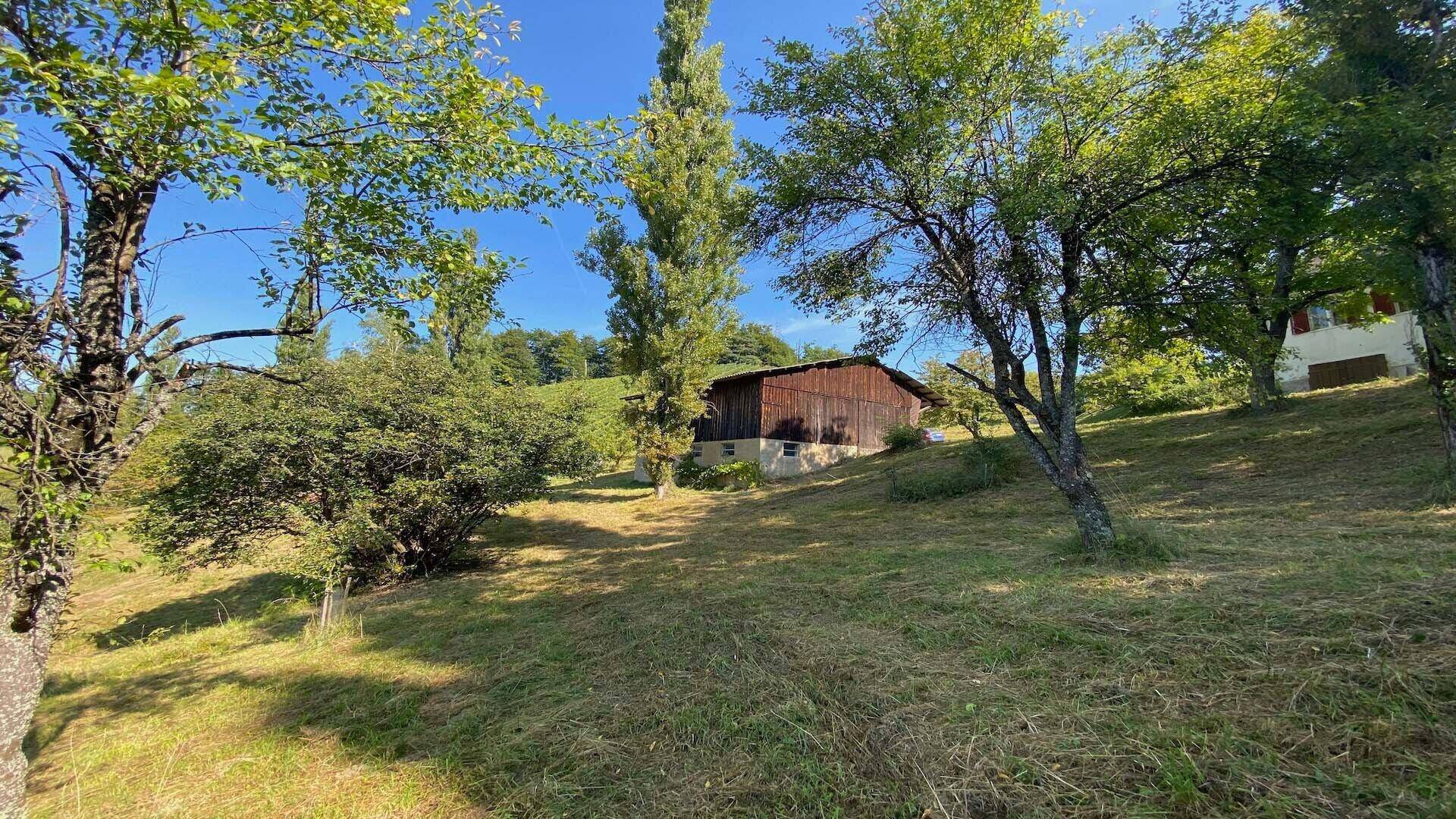Rustikale Scheune auf einem grünen Hügel mit Bäumen und einem Wohnhaus im Hintergrund.