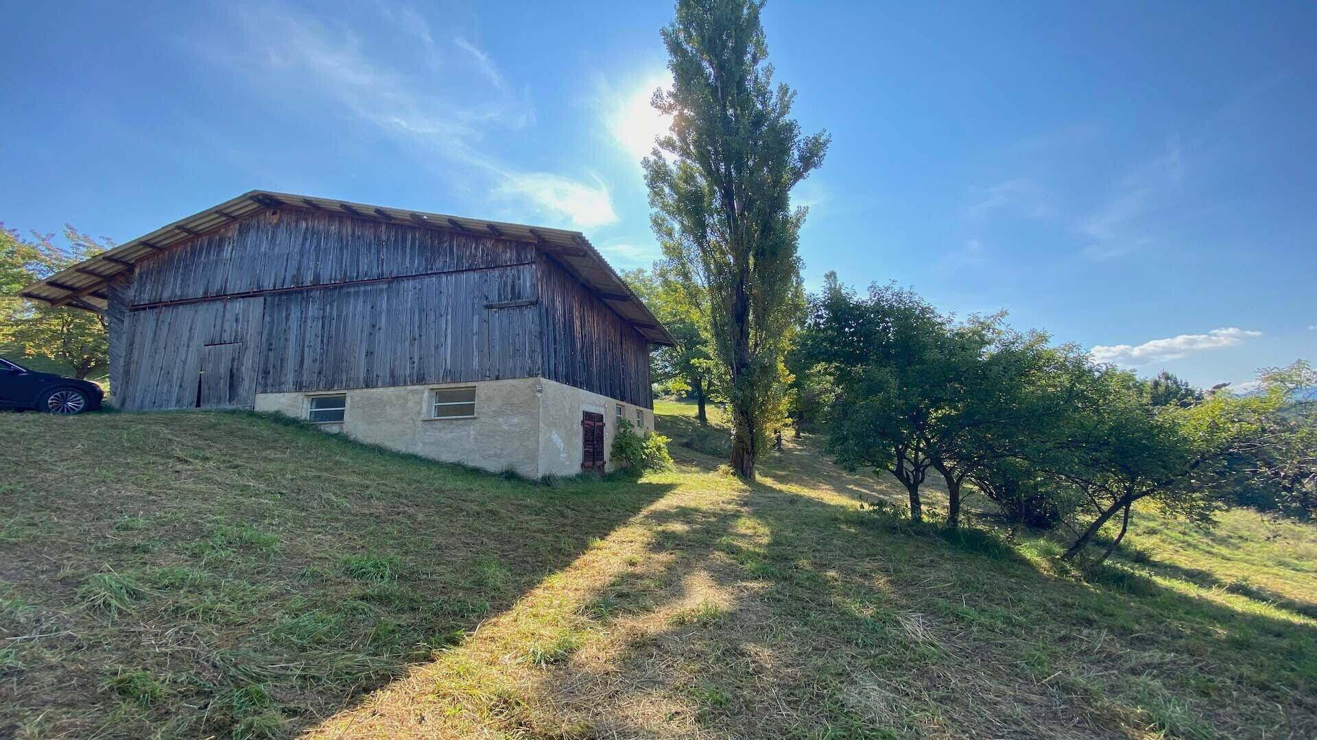 Rustikale Holzscheune mit Steinfundament auf einem sonnigen Hügel, umgeben von Bäumen und Gras.