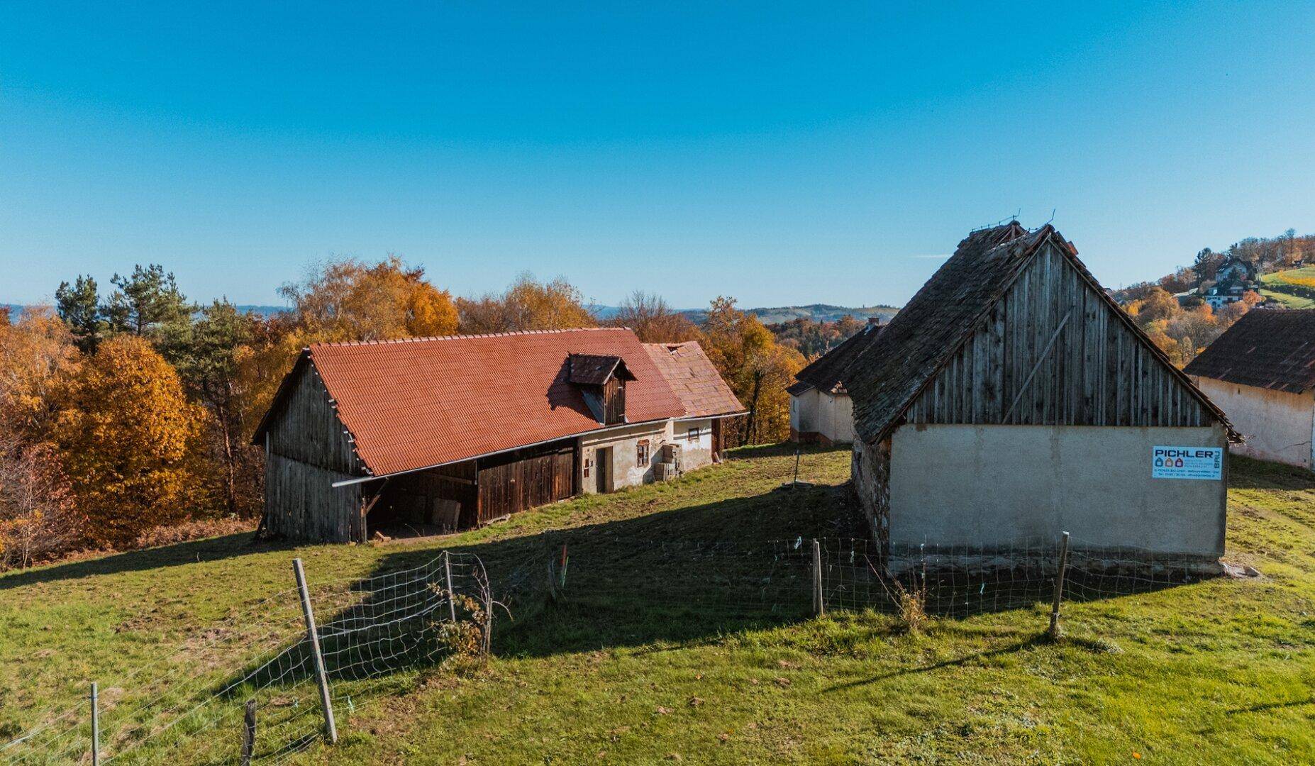 Ländliche Gebäude mit Holzelementen und Ziegeldächern, umgeben von herbstlicher Vegetation und einem Zaun.