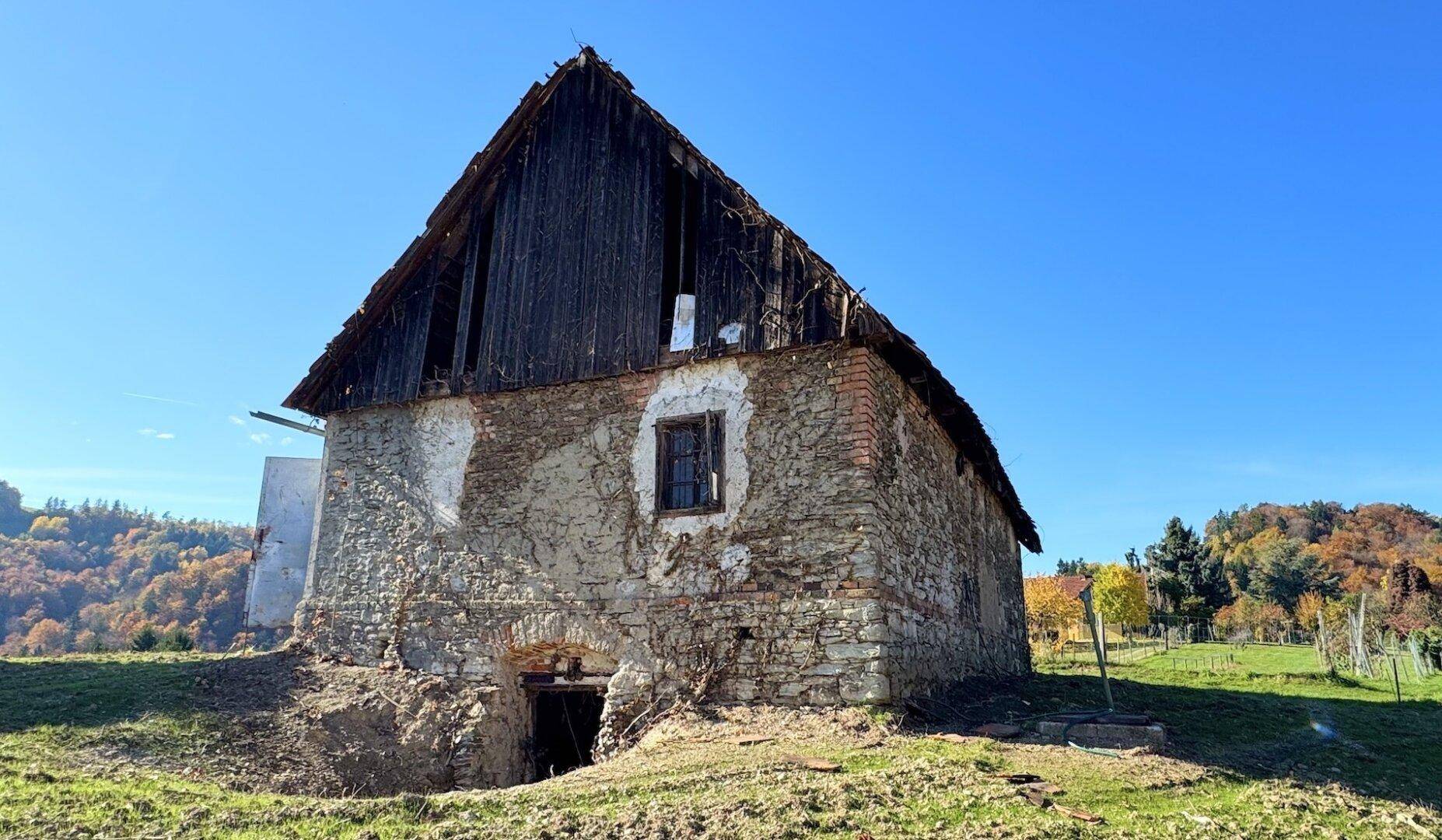 Rustikales Steingebäude mit Holzdach und einem kleinen Kellereingang, umgeben von grüner Landschaft.