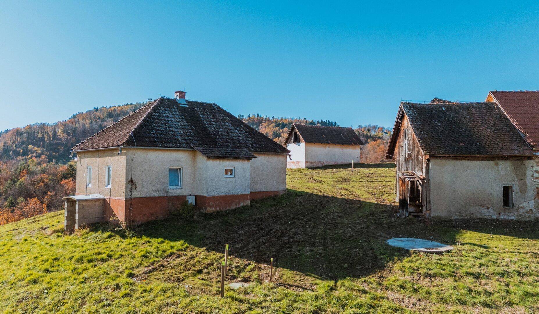 Ländliche Häuser auf einem grünen Hügel mit herbstlicher Vegetation im Hintergrund unter blauem Himmel.