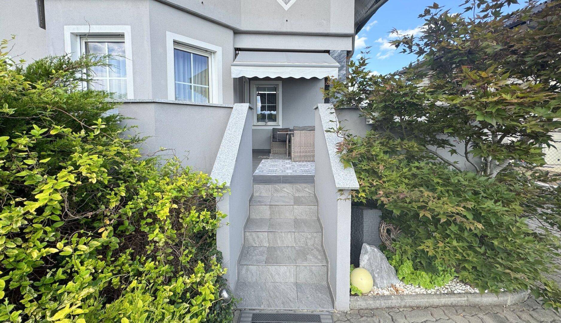 Eingangsbereich des Hauses mit Steintreppe, kleiner Terrasse und grünen Büschen.