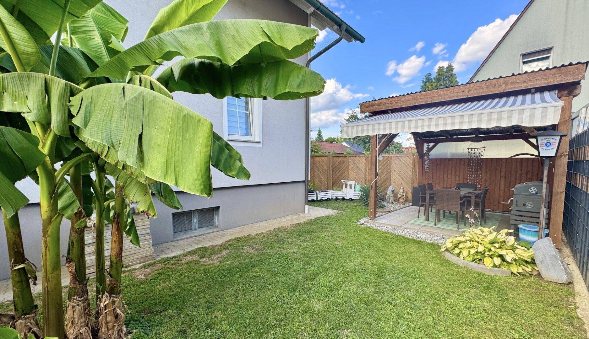 Grüner Gartenbereich mit überdachtem Essplatz, Grill und tropischen Pflanzen.