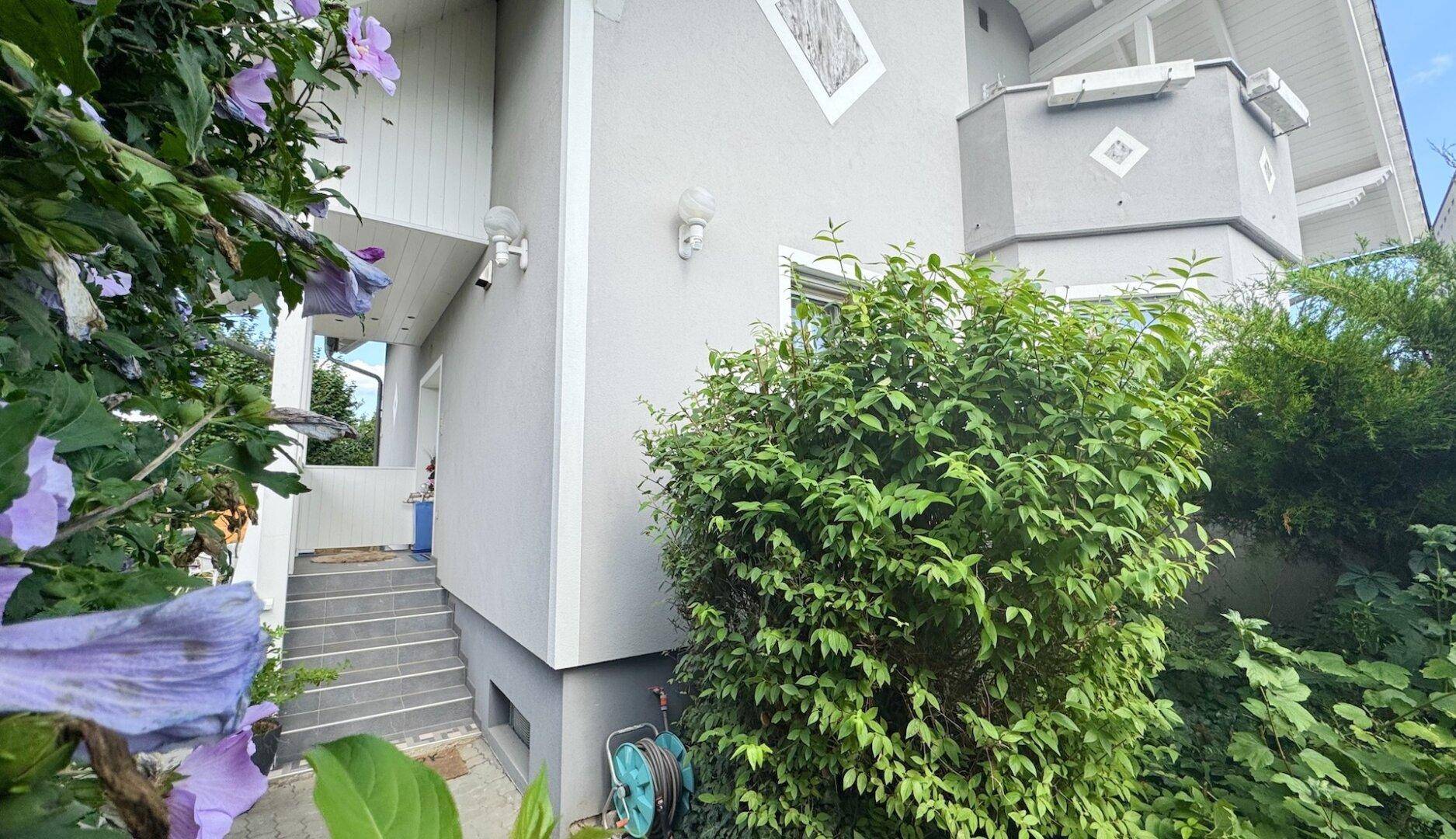Seitlicher Eingangsbereich des Hauses mit Treppe, grünen Sträuchern und blühenden Pflanzen.
