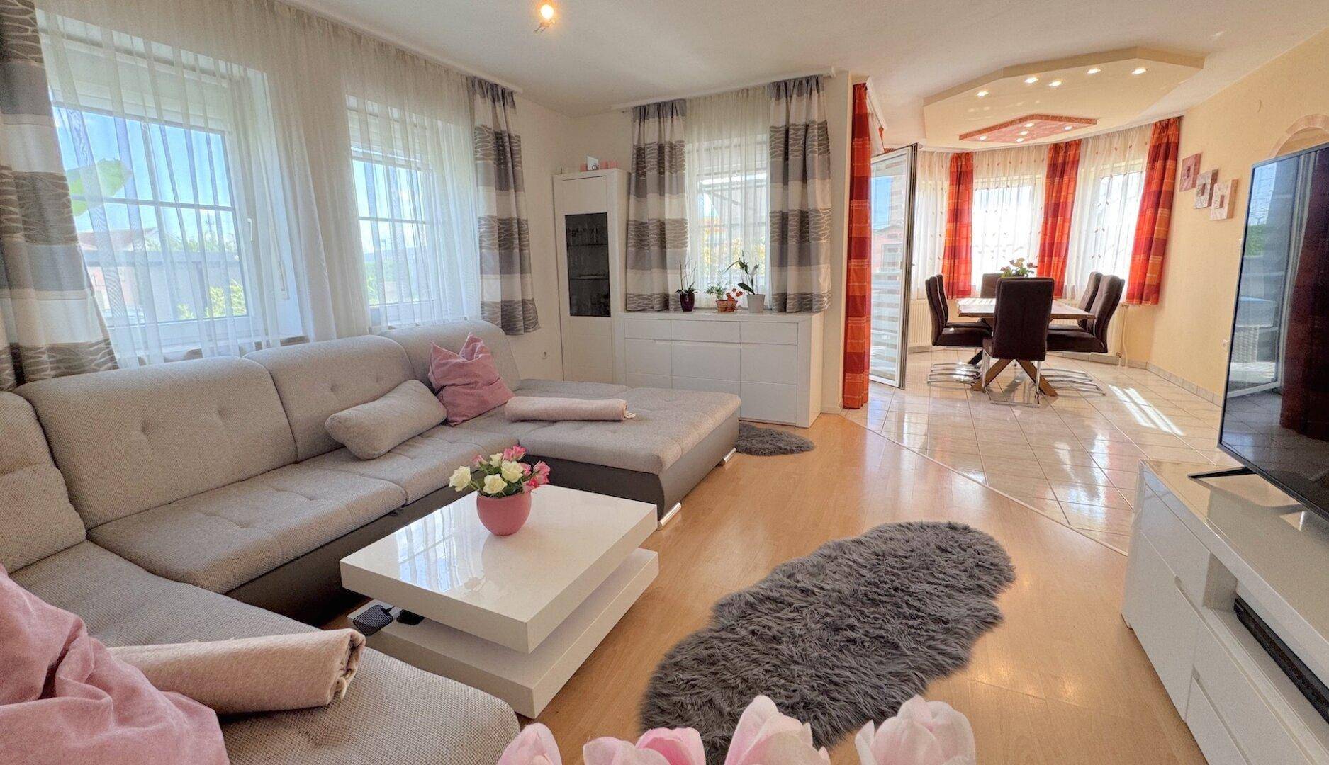 Geräumiger Wohn-Essbereich mit hellem Sofa, Esstisch und großen Fenstern.