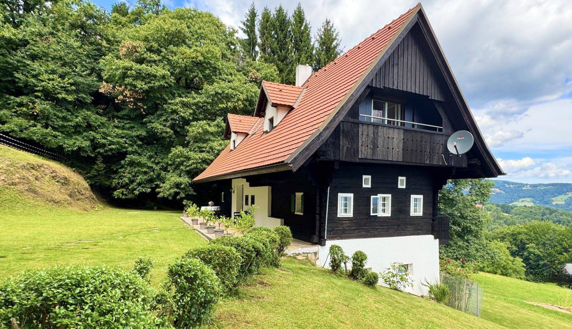 Seitliche Ansicht des Hauses mit Holzbalkon und rotem Ziegeldach, eingebettet in eine grüne Hügellandschaft.