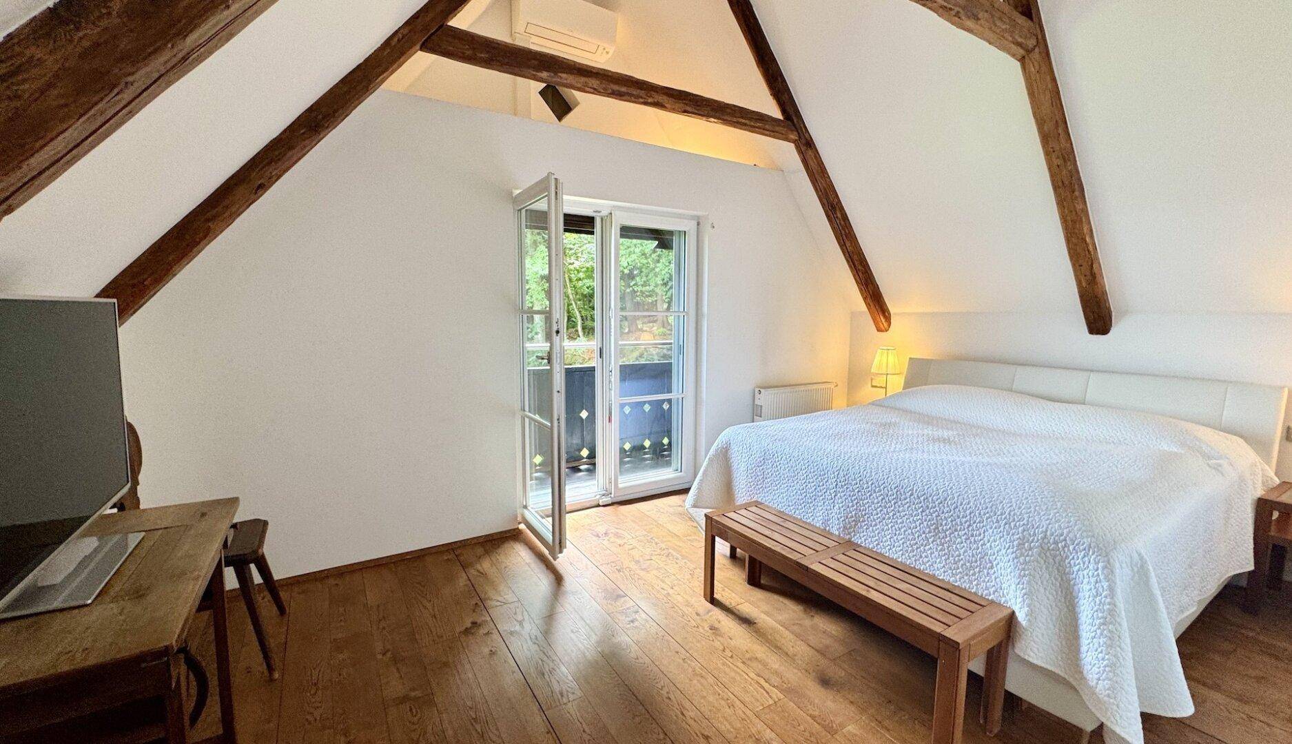 Schlafzimmer im Dachgeschoss mit Holzbalken, Holzboden und Zugang zu einem kleinen Balkon.