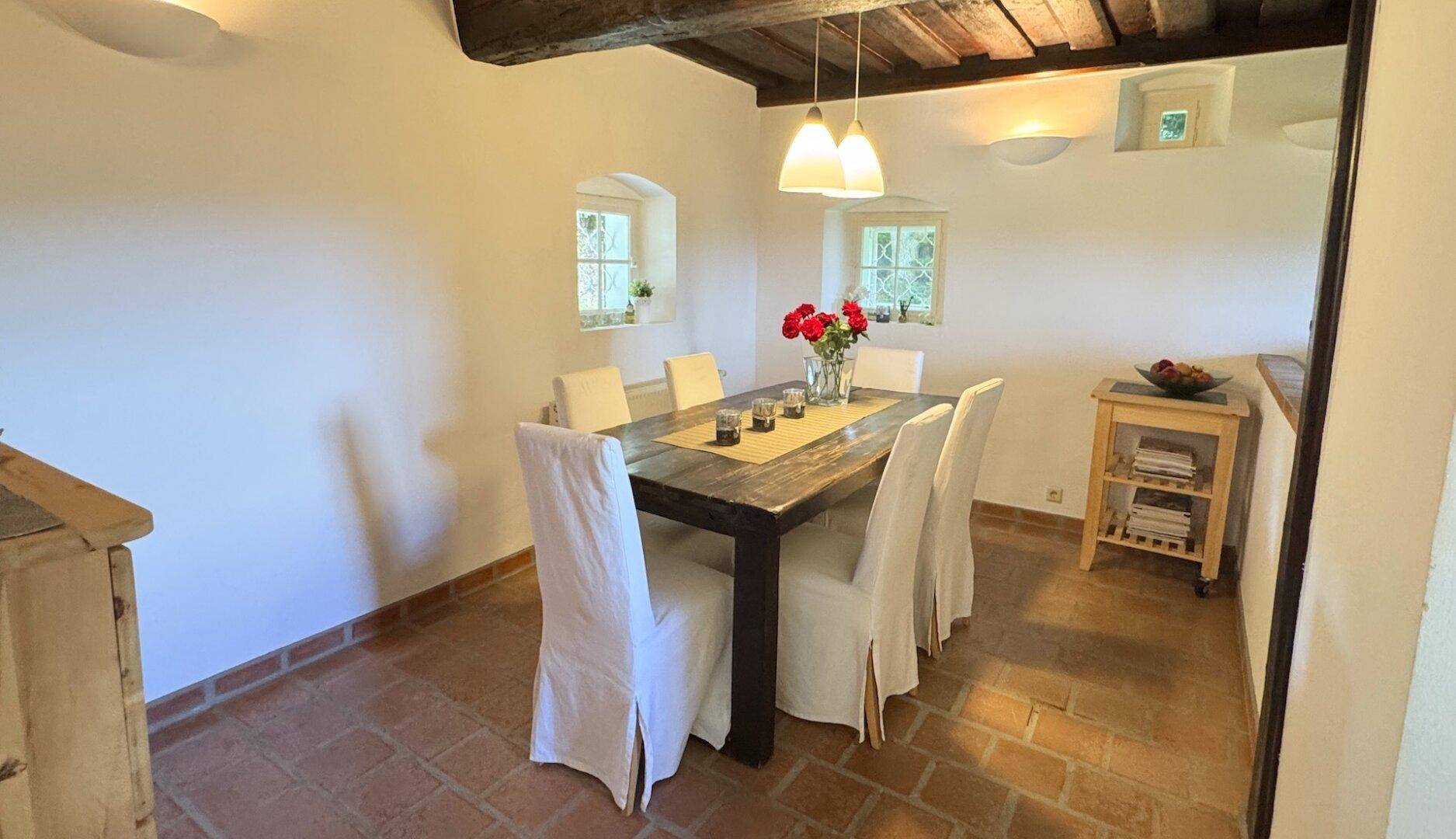 Esszimmer mit rustikalem Holztisch, weißen Stühlen und Terrakottaboden, beleuchtet von Hängelampen.