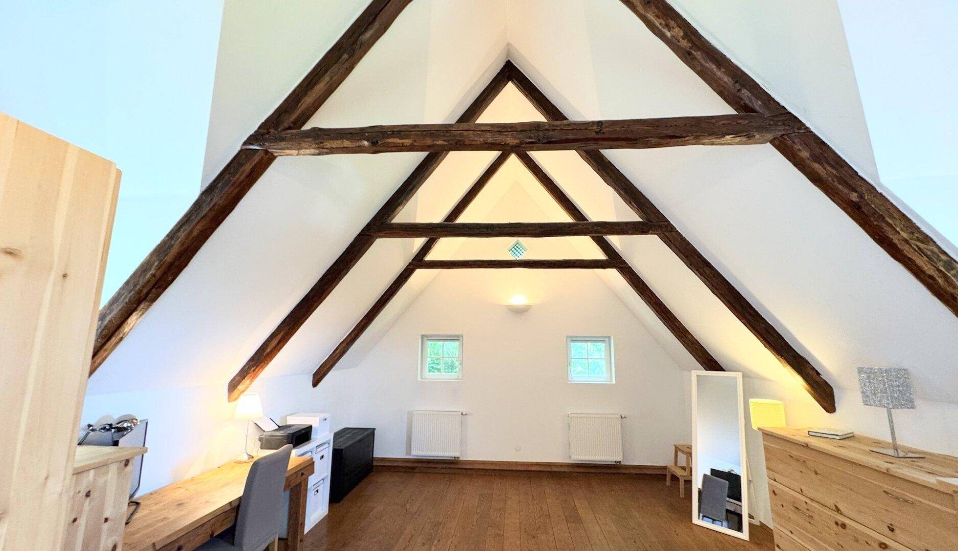 Helles Dachgeschosszimmer mit hohen Decken, Holzbalken und zwei kleinen Fenstern, ideal als Arbeitsbereich.