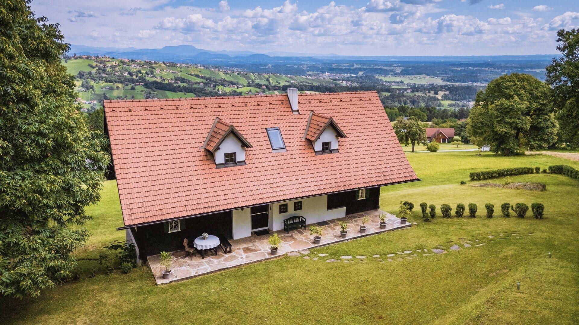 Luftaufnahme des Landhauses mit rotem Ziegeldach, umgeben von grünen Wiesen und einer weiten Landschaft.