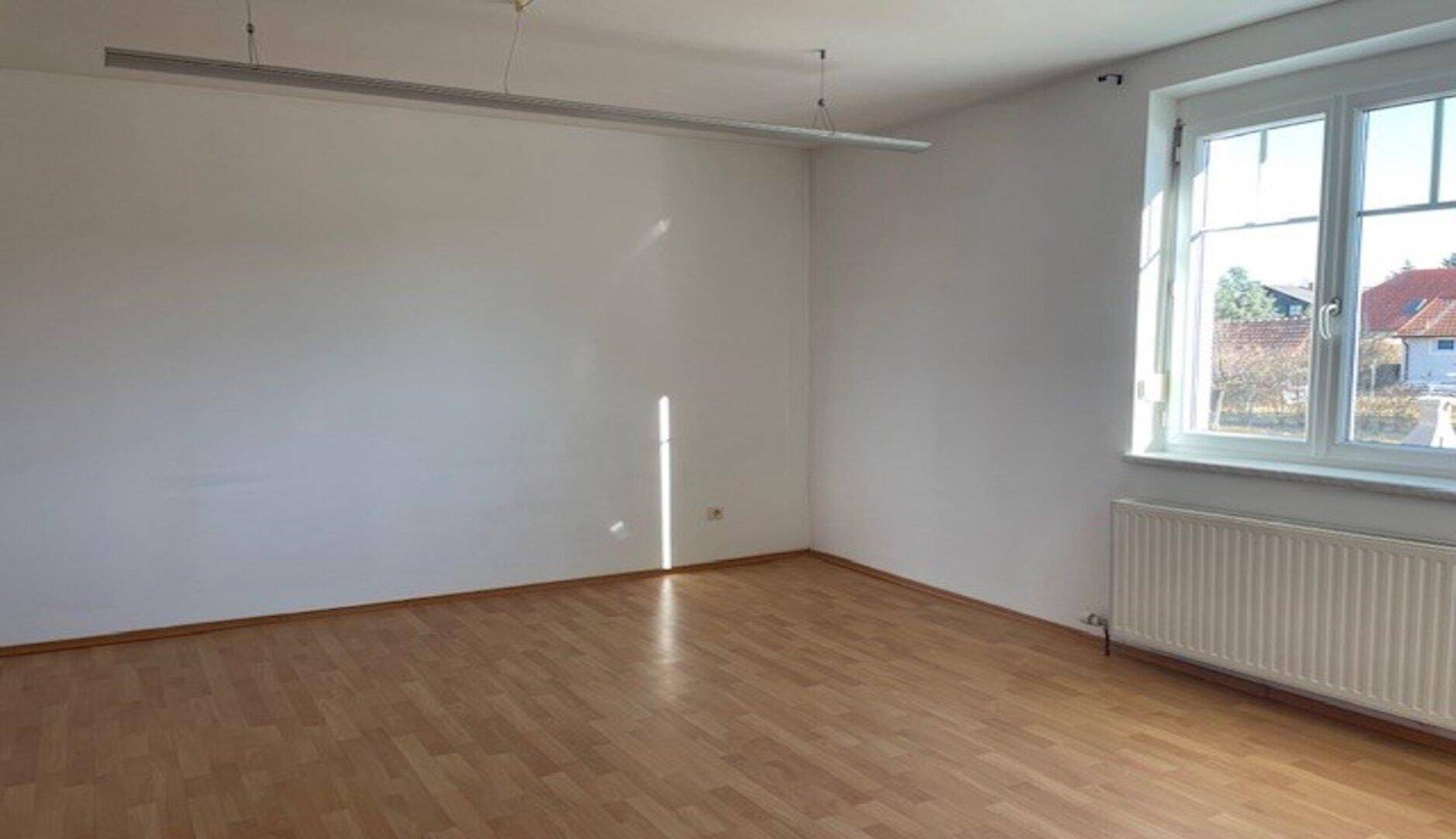 Leerer Raum mit hellem Holzboden, weißen Wänden und einem großen Fenster für viel Tageslicht.
