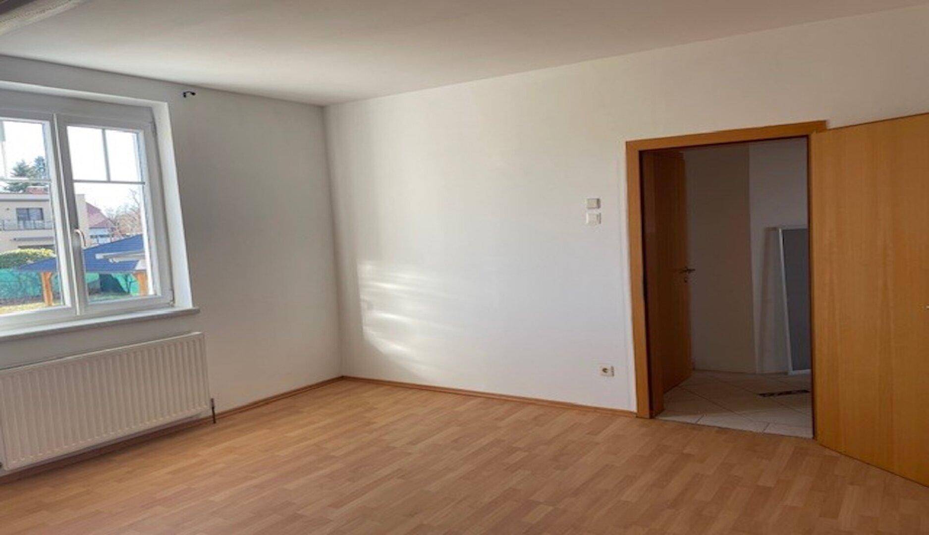 Heller Raum mit Holzboden und weiß gestrichenen Wänden, mit Zugang zu einem weiteren Zimmer.