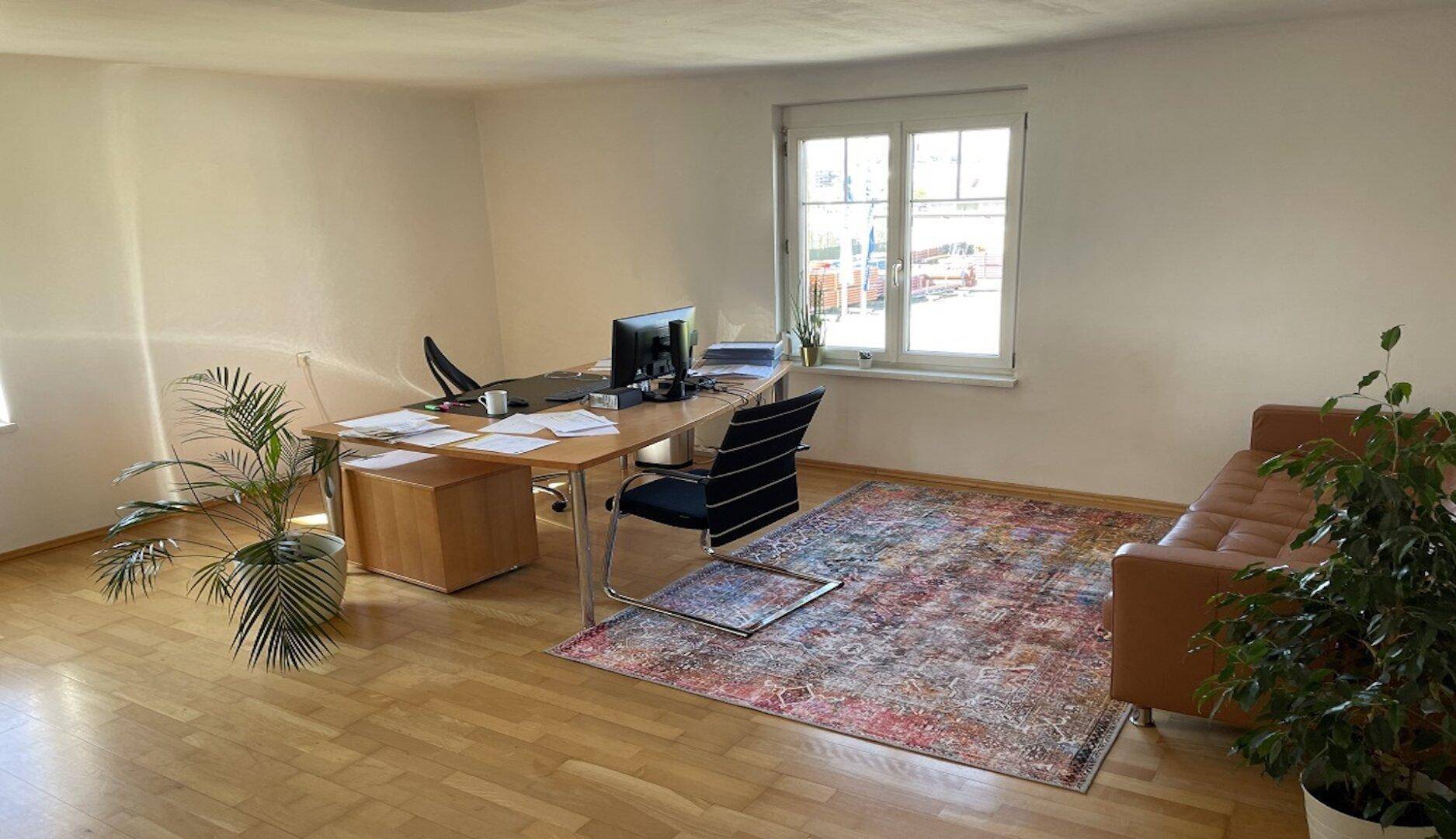 Geräumiges Büro mit Holzboden, Schreibtisch und bequemer Couch, hell durch große Fenster.