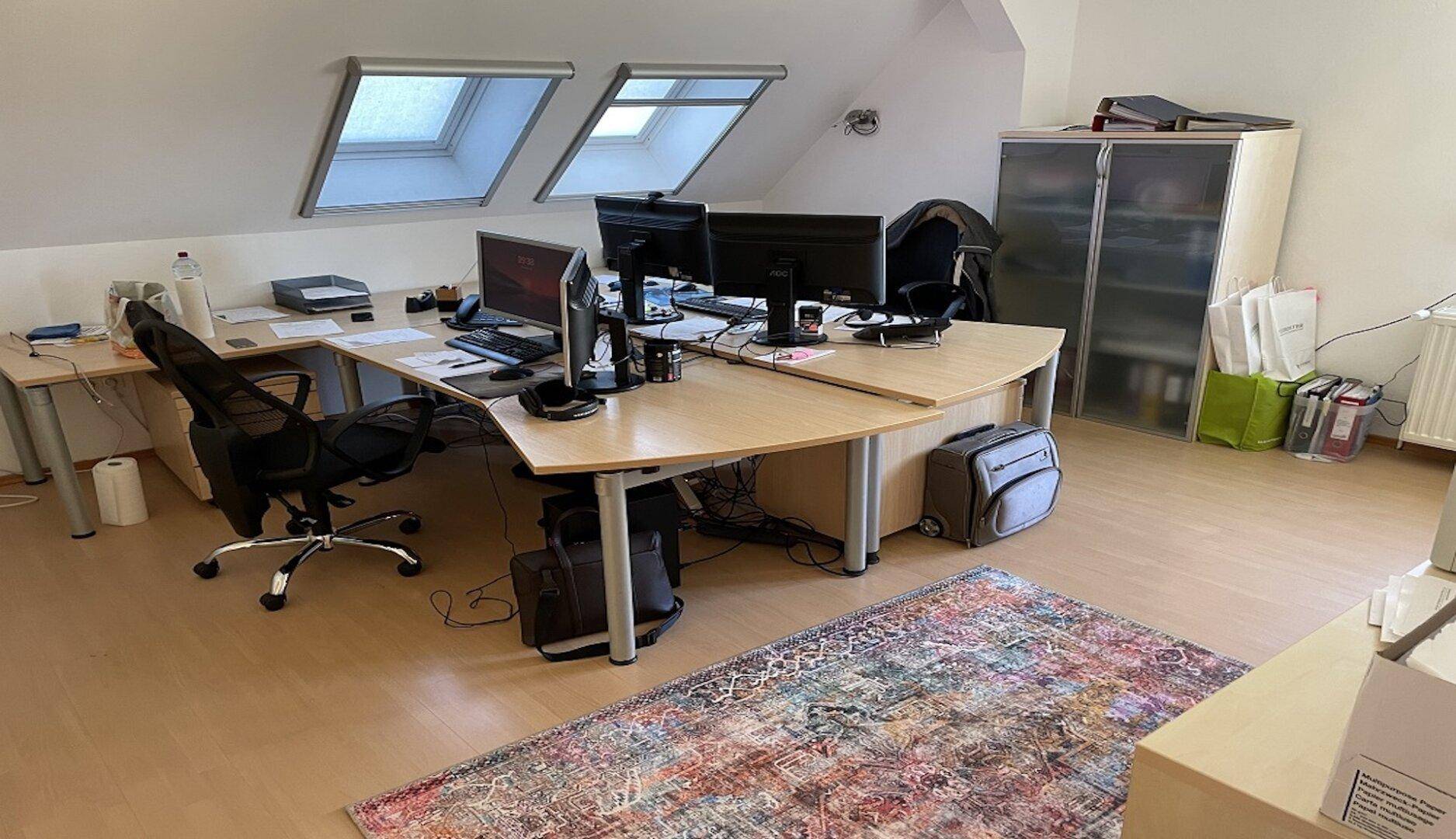 Dachgeschossbüro mit mehreren Schreibtischen, Dachfenstern und Teppichboden, gut beleuchtet.