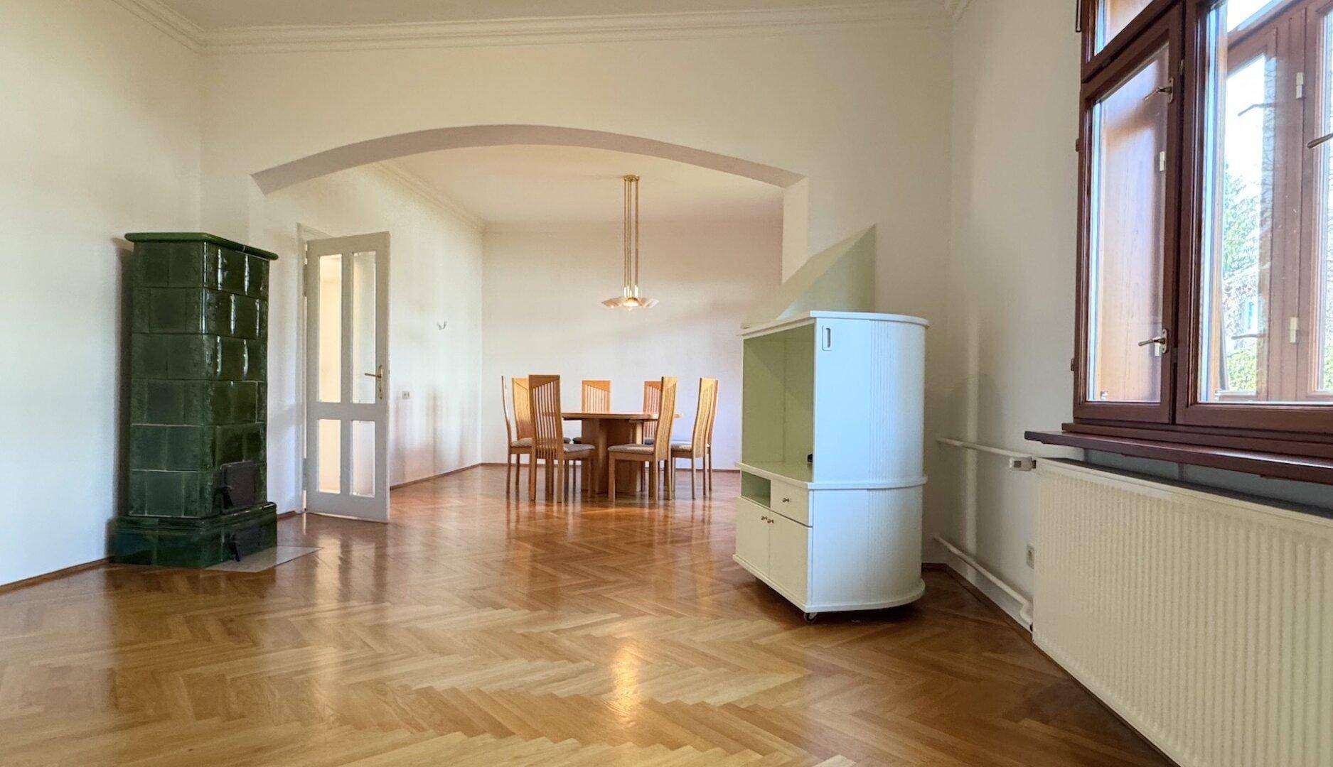 Offener Wohn-Essbereich mit Fischgrätparkett, Kachelofen und einem Esstisch mit Stühlen.